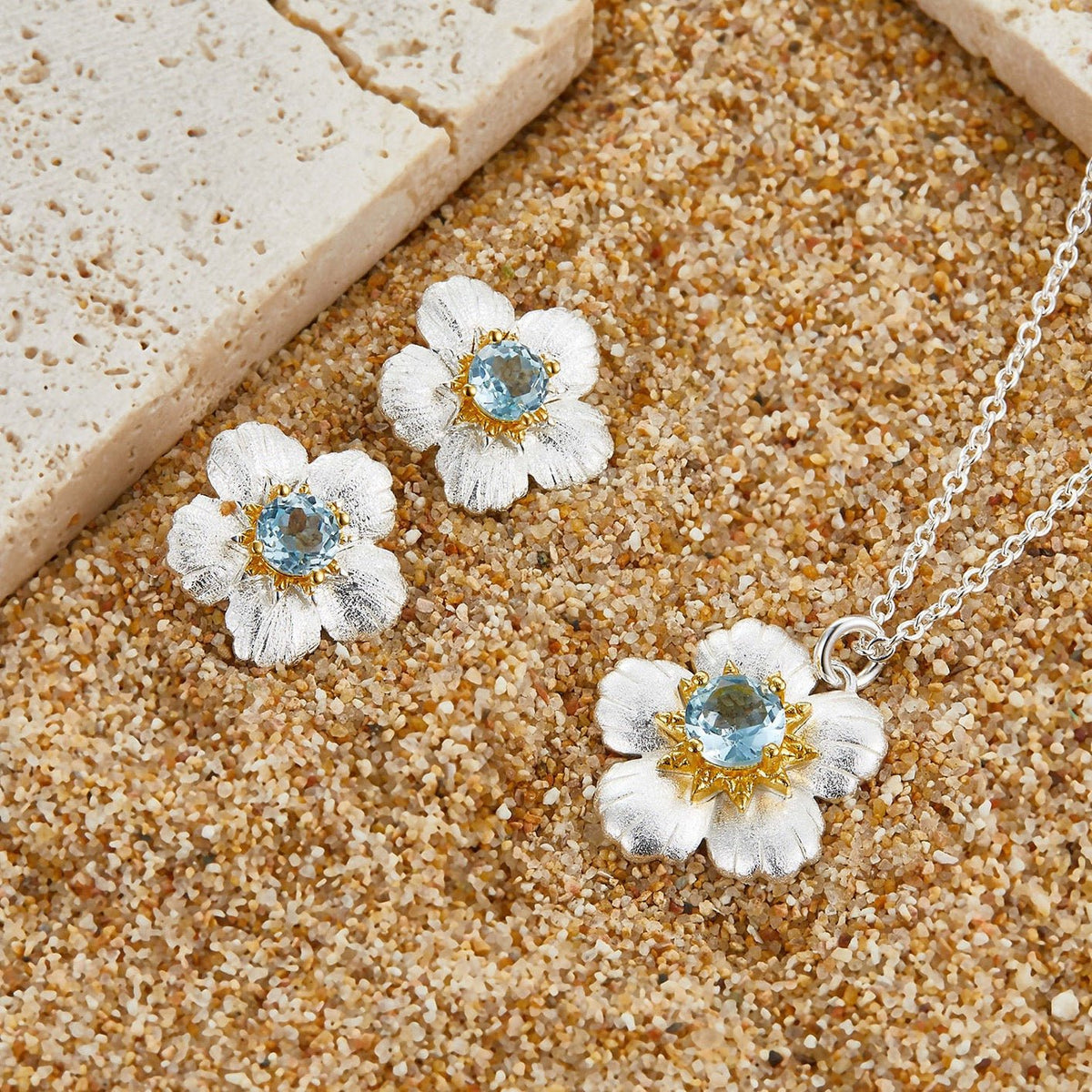 Matching Bleu de Jardin floral jewelry set with blue topaz stud earrings and pendant necklace laid on a textured surface.