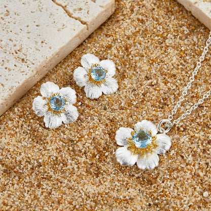 Matching Bleu de Jardin floral jewelry set with blue topaz stud earrings and pendant necklace laid on a textured surface.